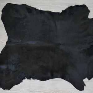 Puede incluir: Una alfombra grande de piel de vaca negra de forma irregular. La piel tiene una superficie suave y texturizada con un ligero brillo. Los bordes son irregulares, mostrando la forma natural de la piel. La alfombra está sobre un suelo de madera clara.