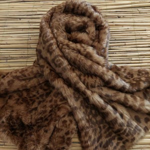 May include: A brown faux fur scarf with a leopard print pattern. The scarf is draped over a bamboo mat, showcasing its soft texture and warm color tones. The design features dark brown spots on a lighter brown background.