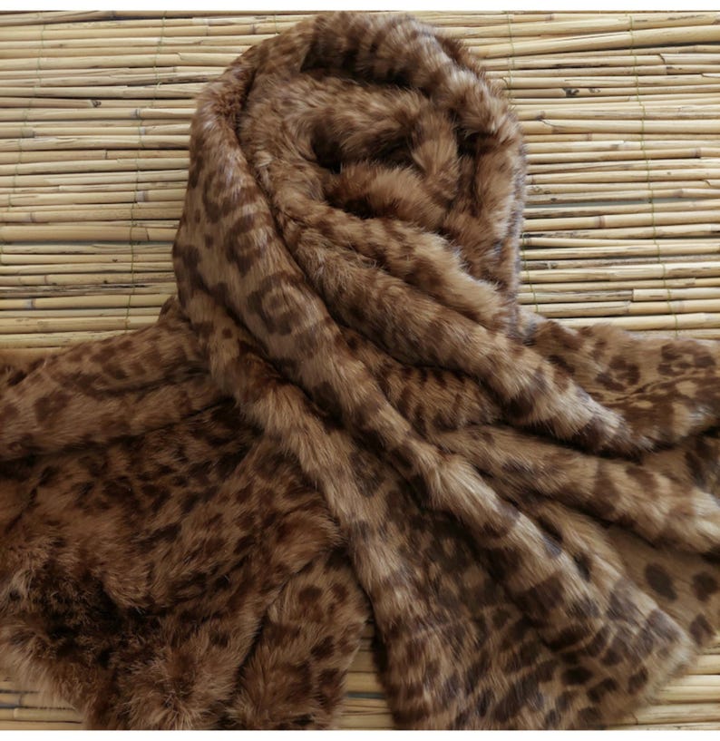 May include: A brown faux fur scarf with a leopard print pattern. The scarf is draped over a bamboo mat, showcasing its soft texture and warm color tones. The design features dark brown spots on a lighter brown background.