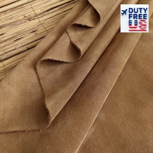 Può includere: Primo piano di un pezzo di tessuto scamosciato marrone piegato. Il tessuto ha una superficie morbida e strutturata ed è drappeggiato in pieghe sciolte. I bordi del tessuto sono leggermente sfilacciati. Un logo "DUTY FREE US" si trova nell'angolo in alto a destra.