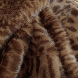 May include: Close-up of a brown and tan faux fur fabric. The fabric has a pattern of dark brown stripes and spots against a lighter brown background, creating a textured appearance. The material appears soft and plush.