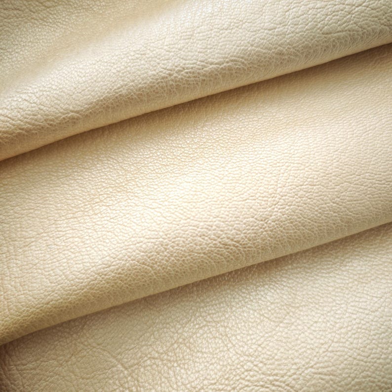 May include: Close-up of a folded piece of cream-colored leather. The leather has a textured surface with a subtle grain pattern. The folds create shadows and highlights, adding depth to the image. The material appears soft and supple.