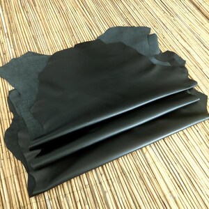 May include: Several sheets of black leather are layered on top of each other, with a few sheets of dark gray leather visible in the background. The leather has a smooth, shiny surface and irregular edges. The background is a light brown, woven material.