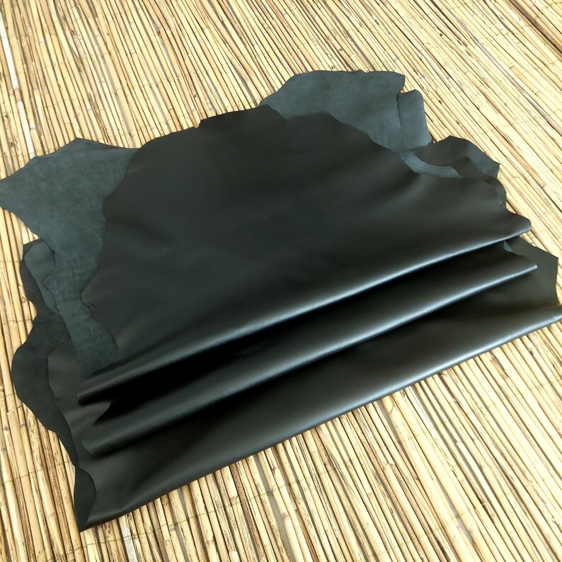 May include: Several sheets of black leather are layered on top of each other, with a few sheets of dark gray leather visible in the background. The leather has a smooth, shiny surface and irregular edges. The background is a light brown, woven material.