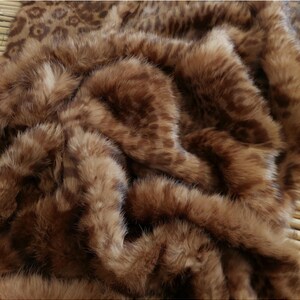 May include: A close-up of a brown faux fur blanket with a leopard print design. The blanket has a soft, plush texture and is folded, creating shadows and depth. The color palette is warm, with shades of brown and tan.