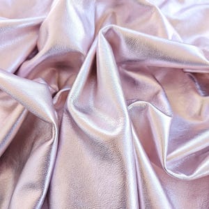 Può includere: Un pezzo di tessuto rosa chiaro con una finitura metallica riflettente e una superficie testurizzata. Il materiale è drappeggiato in pieghe morbide e profonde, creando riflessi e ombre che enfatizzano la qualità lucida e liscia del tessuto.