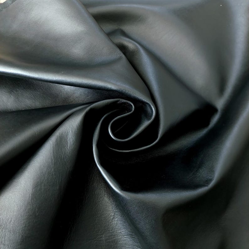 May include: Close-up of a swatch of black leather fabric. The material is smooth and shiny, with subtle variations in texture. The fabric is draped and folded, creating shadows and highlights that emphasize its supple quality. Ideal for upholstery, clothing, or accessories.