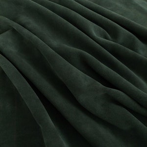 May include: A close-up of a dark green fabric with soft folds and wrinkles. The fabric has a smooth, velvety texture.