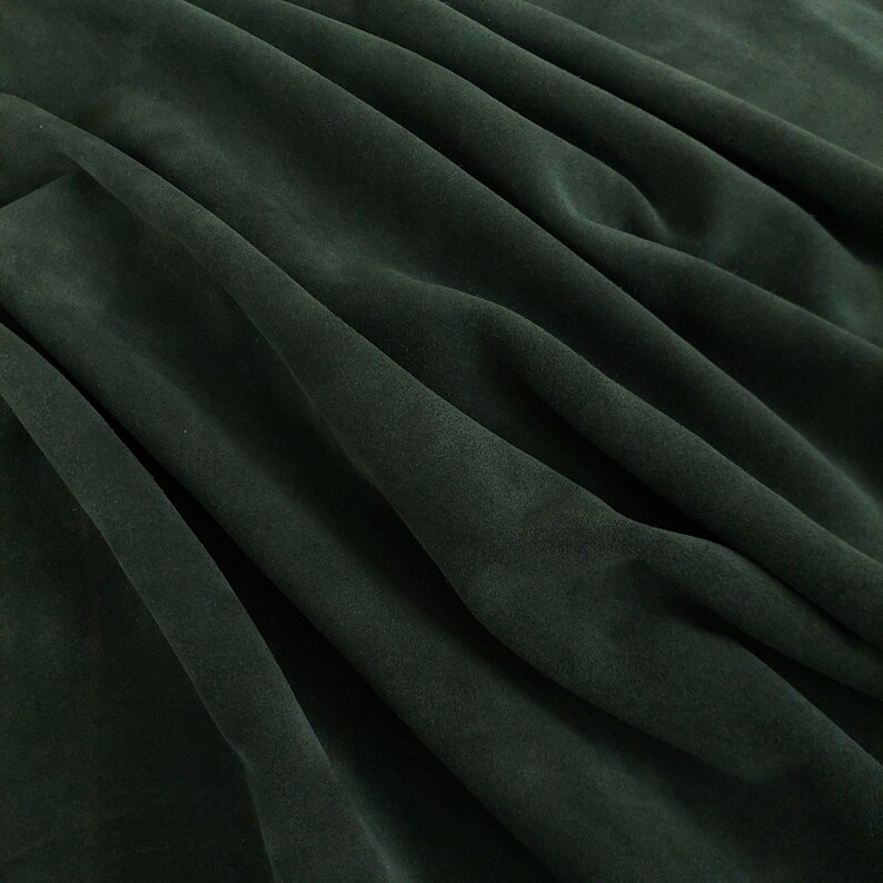 May include: A close-up of a dark green fabric with soft folds and wrinkles. The fabric has a smooth, velvety texture.