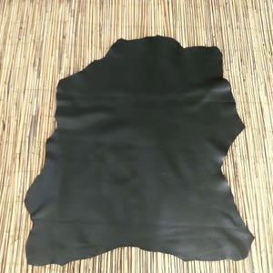 May include: A large, irregularly shaped piece of black leather. The leather has a smooth, matte finish and appears to be a single hide. The background is a light brown, textured surface.
