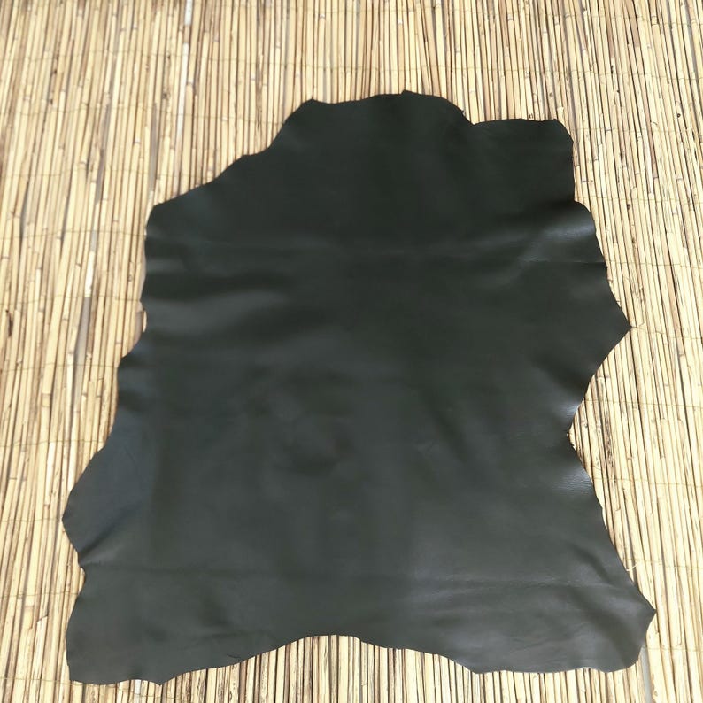 May include: A large, irregularly shaped piece of black leather. The leather has a smooth, matte finish and appears to be a single hide. The background is a light brown, textured surface.