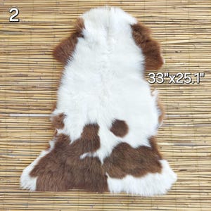 May include: A brown and white fur rug, measuring 33" x 25.1". The rug has a natural, irregular shape with a mix of brown and white fur. The fur appears soft and plush, with a spotted pattern. The rug is displayed on a light brown, textured surface.