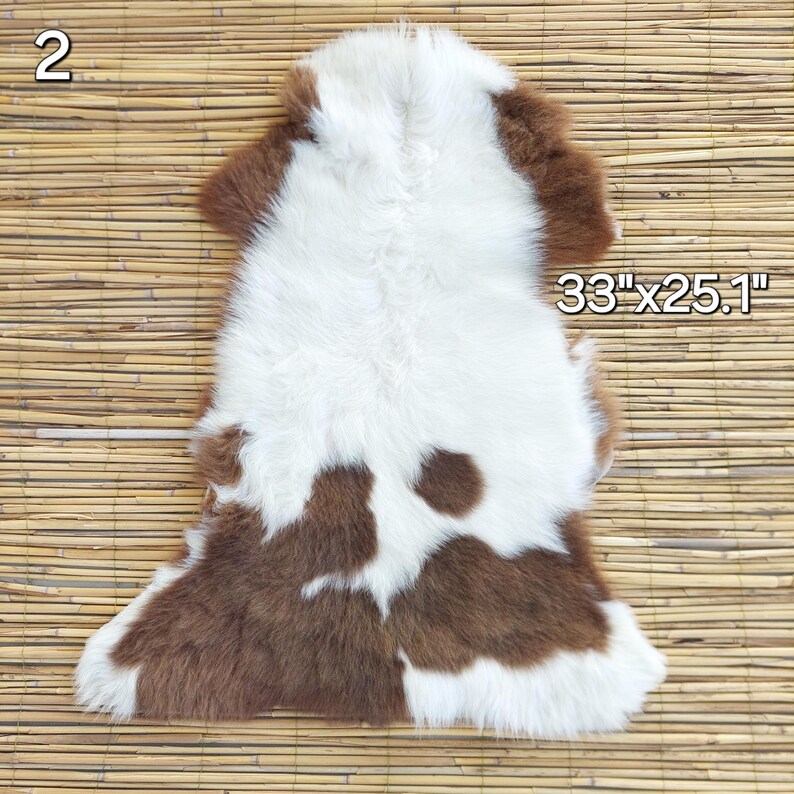 May include: A brown and white fur rug, measuring 33" x 25.1". The rug has a natural, irregular shape with a mix of brown and white fur. The fur appears soft and plush, with a spotted pattern. The rug is displayed on a light brown, textured surface.