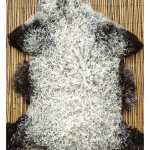 May include: A sheepskin rug with a natural, curly texture. The rug features a mix of white, gray, and dark brown tones. The wool fibers are densely packed, creating a soft and inviting appearance. The rug is laid on a bamboo mat.