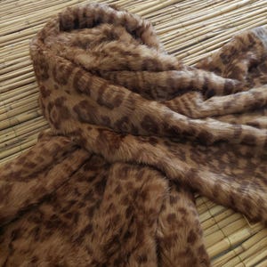 May include: A brown faux fur coat with a hood. The coat features a leopard print pattern in shades of brown and tan. The fabric appears soft and plush, with a close-up view highlighting the texture and design.