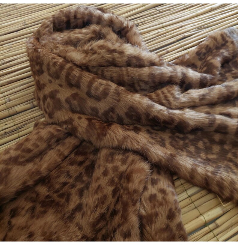 May include: A brown faux fur coat with a hood. The coat features a leopard print pattern in shades of brown and tan. The fabric appears soft and plush, with a close-up view highlighting the texture and design.