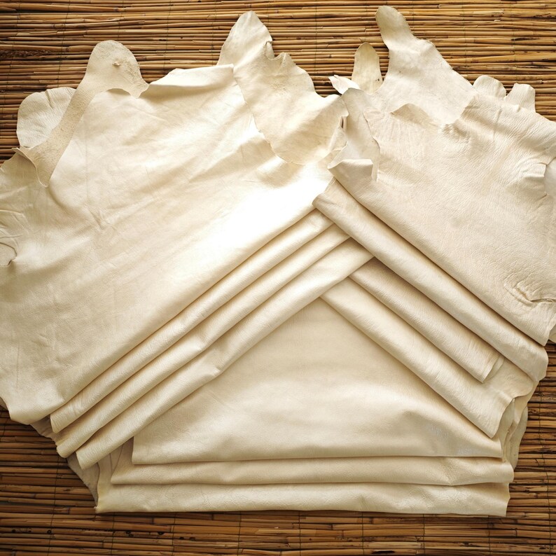 May include: A collection of cream-colored leather hides, some folded and stacked, others spread out. The hides have irregular edges and a slightly textured appearance. The background is a woven bamboo mat.