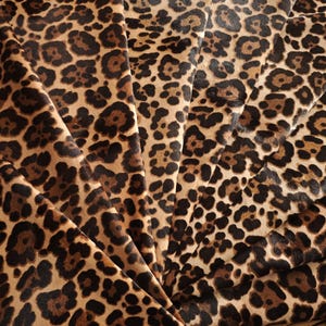 Puede incluir: Primer plano de una tela con estampado de leopardo. La tela tiene un fondo marrón claro con manchas marrones oscuras. La tela está doblada en forma de abanico, mostrando el diseño.