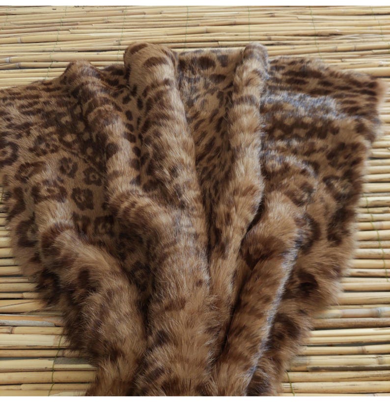 May include: A brown faux fur throw blanket with a leopard print pattern. The blanket is folded, showing the soft texture and varying shades of brown. The throw is displayed on a bamboo mat.