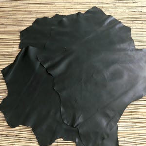 May include: Several sheets of dark gray leather are arranged on a light brown, textured surface. The leather pieces have irregular, wavy edges and a smooth, matte finish. The leather appears to be supple and of high quality, suitable for crafting.