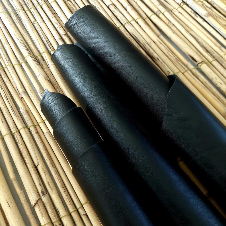 May include: Three rolls of black leather are displayed on a bamboo mat. The leather has a smooth, matte finish and varying sizes. The rolls are tightly wound, showcasing the material's flexibility and texture. The lighting highlights the leather's dark color.