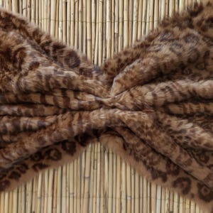 May include: A faux fur throw blanket with a brown and tan leopard print pattern. The blanket is soft and plush, with a textured surface. The background is a bamboo mat.