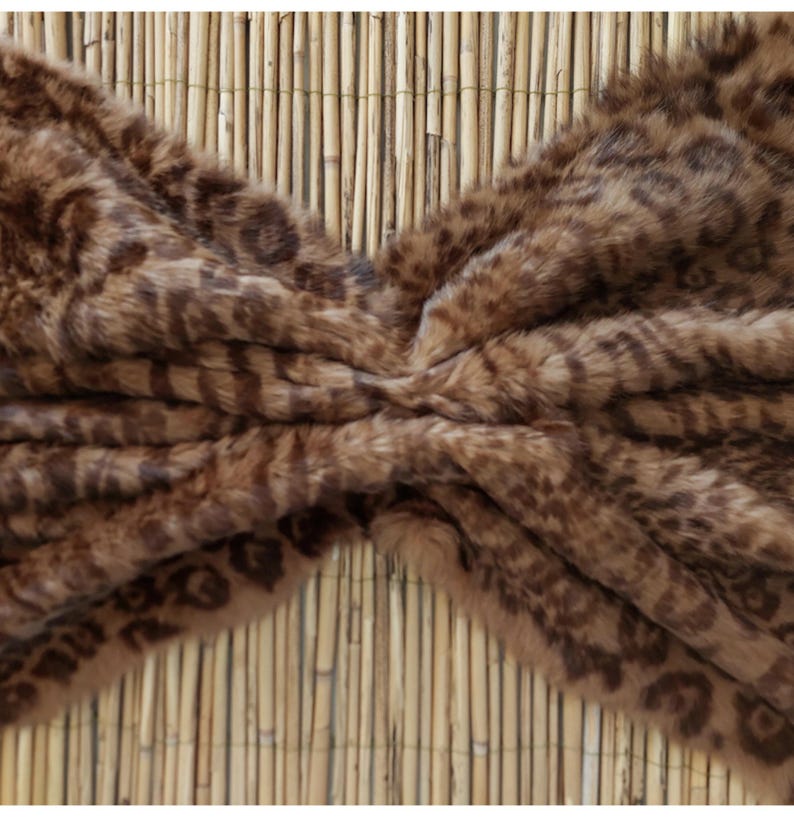 May include: A faux fur throw blanket with a brown and tan leopard print pattern. The blanket is soft and plush, with a textured surface. The background is a bamboo mat.