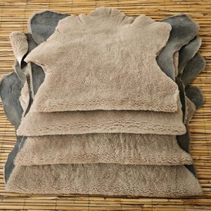 Pode incluir: Uma pilha de tapetes de pele de ovelha em tons de bege e cinza. Os tapetes têm uma superfície macia e texturizada e formas irregulares. O fundo é um tapete de bambu natural.