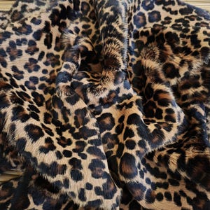 May include: A close-up shot of a leopard print faux fur fabric. The fabric has a tan background with dark brown and black spots. The material looks soft and fluffy, with a realistic animal print design.