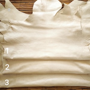 May include: Close-up of several pieces of off-white leather, with a textured surface and irregular edges. The leather is stacked, with the numbers 1, 2, and 3 visible on the edges. The background is a woven bamboo mat.