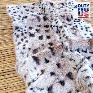 May include: A faux fur throw blanket with a spotted pattern in shades of cream, brown, and black. The blanket has a soft, plush texture and is folded over on a bamboo mat. The text "DUTY FREE" is visible in the upper right corner.