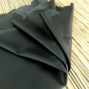 May include: A large piece of black leather, folded and draped, showcasing its smooth, shiny surface. The leather is a deep, solid black, with visible folds and creases. The background is a natural bamboo mat.