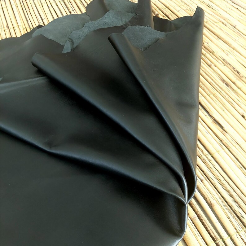 May include: A large piece of black leather, folded and draped, showcasing its smooth, shiny surface. The leather is a deep, solid black, with visible folds and creases. The background is a natural bamboo mat.