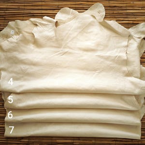 May include: A stack of cream-colored leather hides, with visible textures and folds. The edges of the hides are uneven, and the numbers 4, 5, 6, and 7 are visible on the side. The hides are stacked on a bamboo mat.