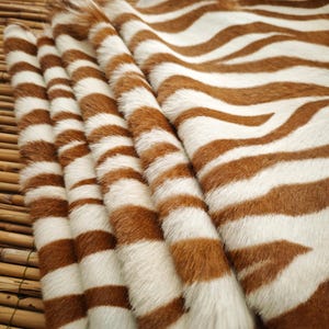 May include: Close-up of a zebra-striped hide. The hide features alternating bands of brown and white, with a soft, textured appearance. The pattern is consistent across the surface, creating a natural, animalistic aesthetic. The material is folded, showing the depth and texture.