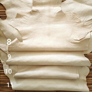 May include: A stack of cream-colored leather hides. The hides are arranged in layers, with the top hide partially unfolded, revealing the textured surface. Numbers 8, 9, 10, and 11 are visible on the left side of the image. The background is a woven bamboo mat.