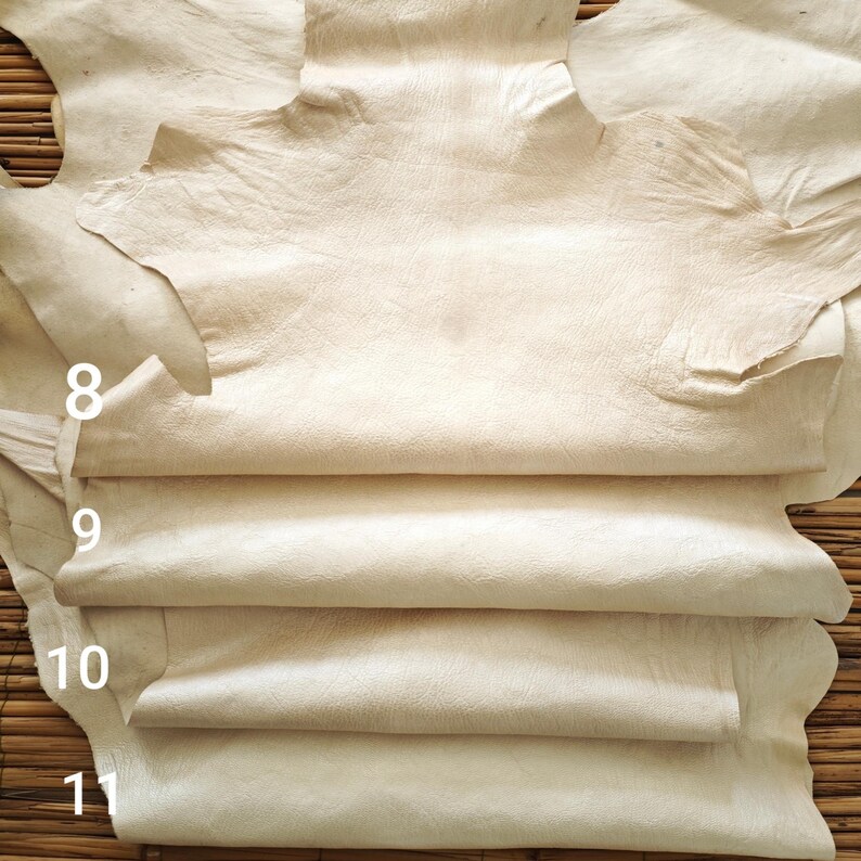 May include: A stack of cream-colored leather hides. The hides are arranged in layers, with the top hide partially unfolded, revealing the textured surface. Numbers 8, 9, 10, and 11 are visible on the left side of the image. The background is a woven bamboo mat.