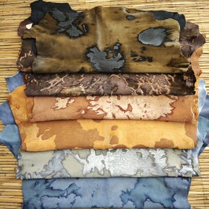 May include: A stack of dyed leather hides in various colors. The hides range from dark brown to light tan, with unique patterns and textures. Some hides have blue, gray, and black accents, creating a natural, organic aesthetic. The hides are stacked on a bamboo mat.
