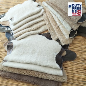 Puede incluir: Una colección de pieles de oveja en tonos crema, beige y marrón. Las pieles tienen un aspecto suave y texturizado, con fibras de lana visibles. Algunas pieles tienen un reverso de cuero marrón oscuro. La imagen incluye un logotipo "DUTY FREE US".