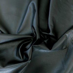 May include: A close-up shot of a piece of black leather fabric. The material is folded and draped, creating shadows and highlights that emphasize its texture and sheen. The leather appears smooth and supple.