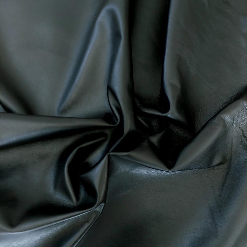 May include: A close-up shot of a piece of black leather fabric. The material is folded and draped, creating shadows and highlights that emphasize its texture and sheen. The leather appears smooth and supple.