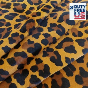 Puede incluir: Primer plano de una tela texturizada con estampado de leopardo. La tela presenta un fondo marrón con manchas marrones oscuras y negras. El logotipo Duty Free US está en la esquina superior derecha.