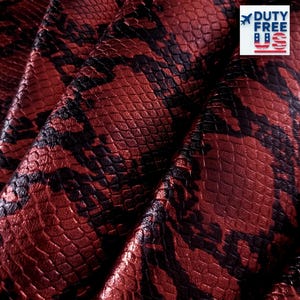 Può includere: Primo piano di un motivo a pelle di serpente rosso e nero strutturato. Il materiale ha una finitura lucida ed è piegato, mostrando le intricate squame e le variazioni di colore. Il testo "DUTY FREE US" è visibile nell'angolo in alto a destra.