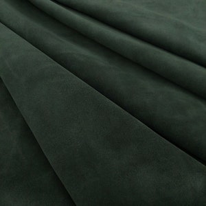 May include: Close-up of a dark green suede fabric with a soft, textured surface. The fabric is folded and draped, showcasing its smooth and luxurious feel.
