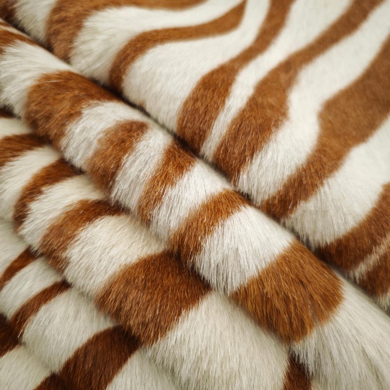 May include: Close-up of a faux fur fabric with a zebra stripe pattern. The fabric features alternating bands of brown and white, creating a textured and visually striking design. The fur appears soft and plush, with visible fibers.