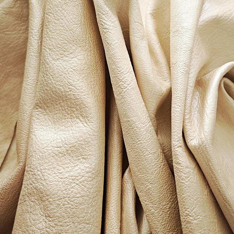 May include: Close-up of beige leather fabric with a textured surface. The leather is draped in soft folds, creating shadows and highlights. The material has a subtle sheen, suggesting a high-quality finish. Ideal for upholstery or crafting.
