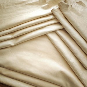 May include: Close-up of several sheets of beige leather, folded and layered. The leather has a smooth, slightly shiny surface with subtle variations in color and texture. The folds create shadows and depth, highlighting the material's flexibility.