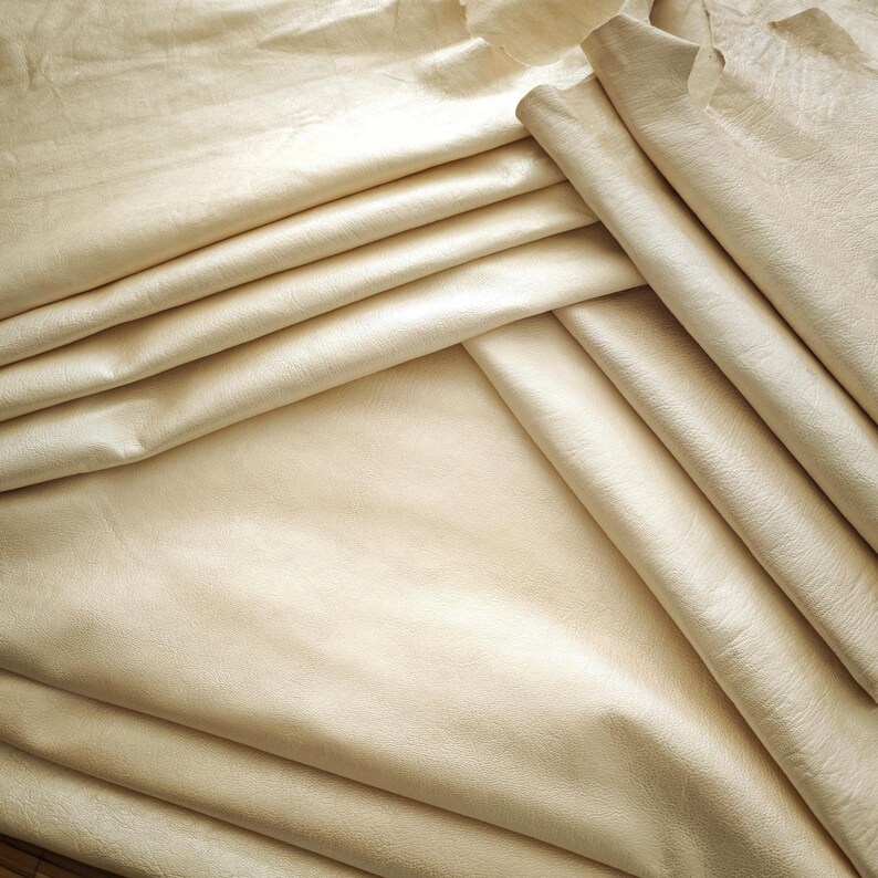 May include: Close-up of several sheets of beige leather, folded and layered. The leather has a smooth, slightly shiny surface with subtle variations in color and texture. The folds create shadows and depth, highlighting the material's flexibility.