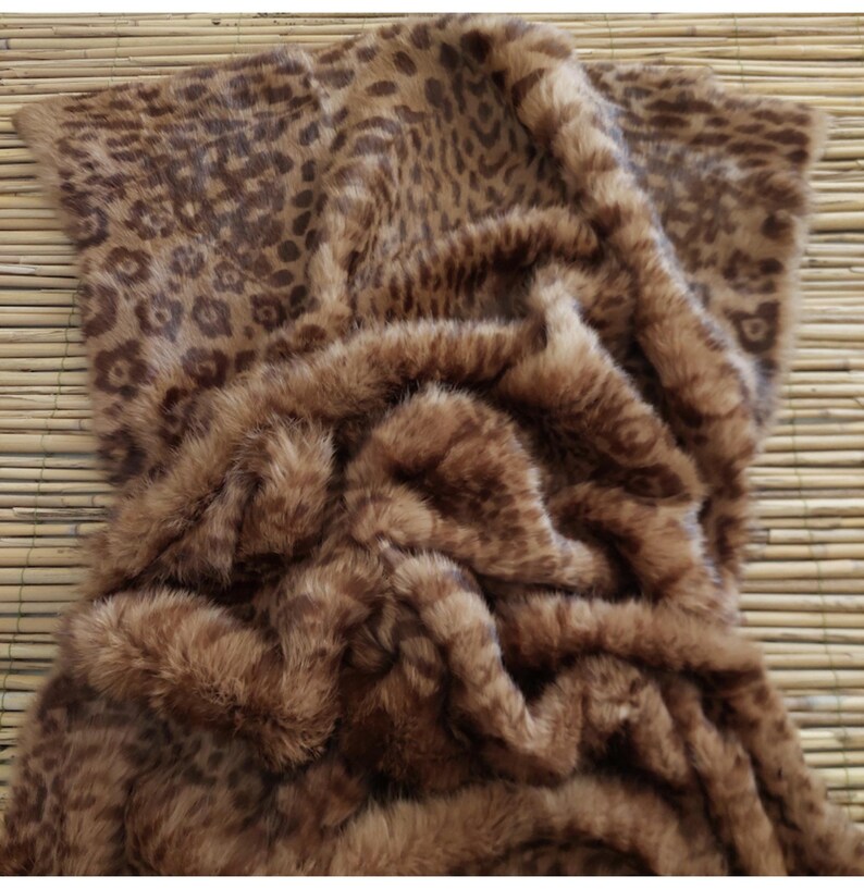 May include: A brown and tan faux fur throw blanket with a leopard print pattern. The blanket is folded and draped, showcasing the soft texture and color variations. The design features spots of varying sizes and shades.