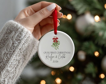 May include: A round white ceramic ornament featuring a red ribbon hanger. The design includes a green mistletoe illustration with a red bow and the text: OUR FIRST CHRISTMAS, Kailyn & Caleb, 2026. The ornament has a smooth, glossy finish.
